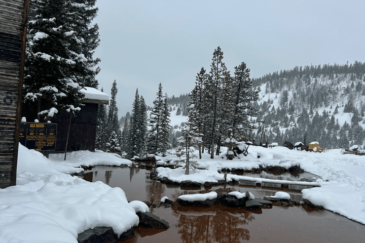 Snowy landscape with trees and a small pond, adjacent to a wooden building.