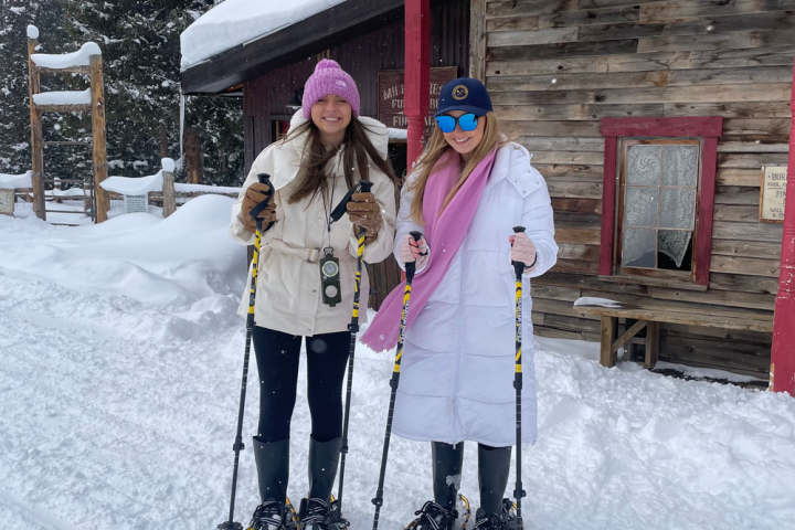 Two women snowshoeing by a snowy cabin, wearing winter clothes and smiling.