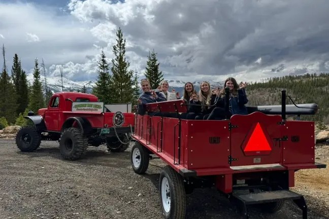 Group sitting in a red wagon near a vintage red truck, with a forested mountain backdrop.