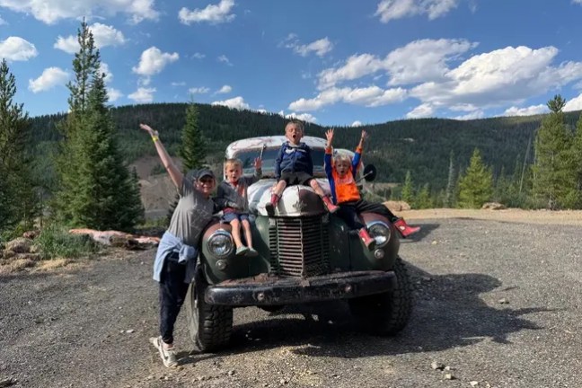 Family on vintage truck in scenic mountain area with trees and blue sky.